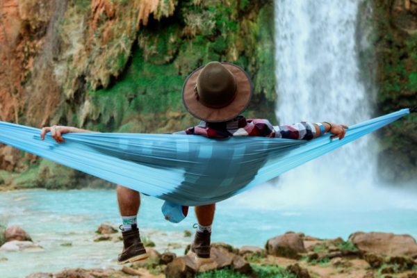 A wide shot of a male lying on a hammock beside a waterfall flowing down from a hill